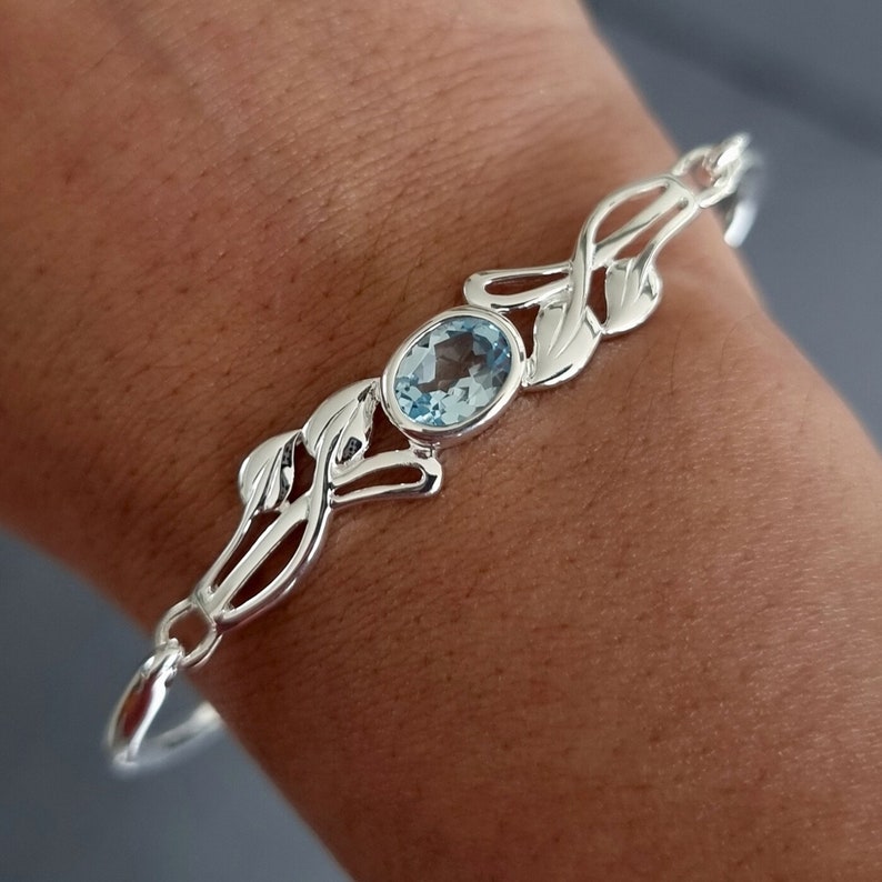 May include: A silver bracelet featuring a central oval-cut light blue gemstone. The metal band is designed with symmetrical, openwork leaf-like motifs extending from the stone setting, creating a continuous, polished metallic link structure.