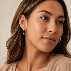 Puede incluir: Un par de pequeños aros de plata pulida con un perfil liso y redondeado. Los pendientes presentan un diseño circular sencillo que se asienta cerca del lóbulo de la oreja, mostrando un acabado metálico reflectante que capta la luz.