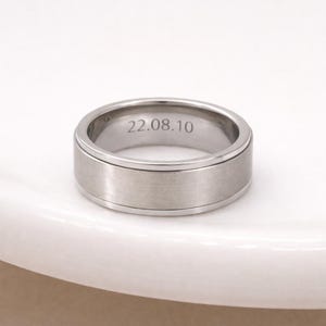 Könnte beinhalten: Ein silberfarbener Ring mit gebürsteter Metalloberfläche. Der Ring hat ein flaches, breites Band mit erhabenem Außenrand. Das Datum "22.08.10" ist auf der Innenseite des Rings eingraviert. Der Ring wird auf einer weißen Oberfläche präsentiert.