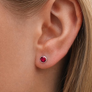 Puede incluir: Primer plano de una oreja con un pequeño pendiente de botón redondo de color rojo rubí engastado en un bisel plateado. El pendiente está centrado en el lóbulo, mostrando el color vibrante y el diseño sencillo de la joya.