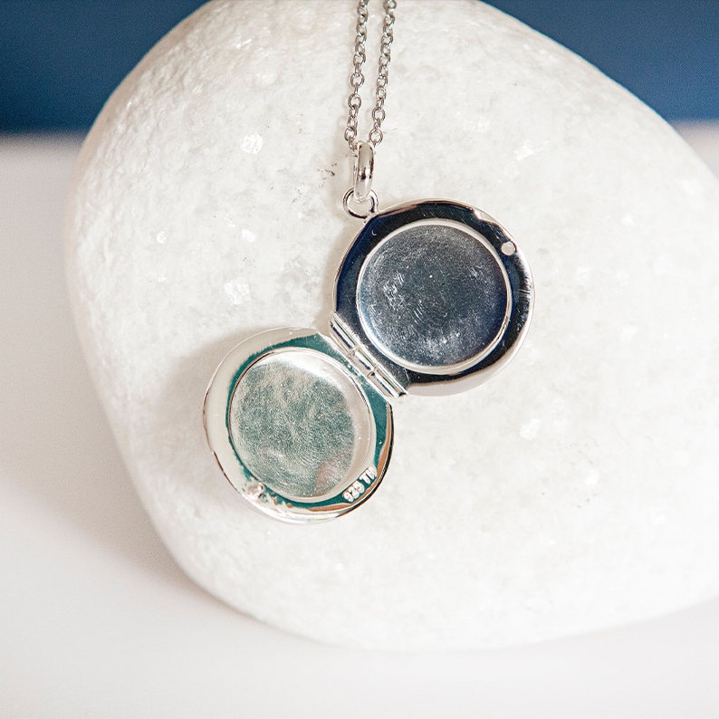 Puede incluir: Un collar con un medall&oacute;n plateado, de forma redonda, abierto para revelar dos compartimentos. El colgante cuelga de una delicada cadena. El medall&oacute;n se muestra sobre una superficie blanca y texturizada. Una joya cl&aacute;sica.