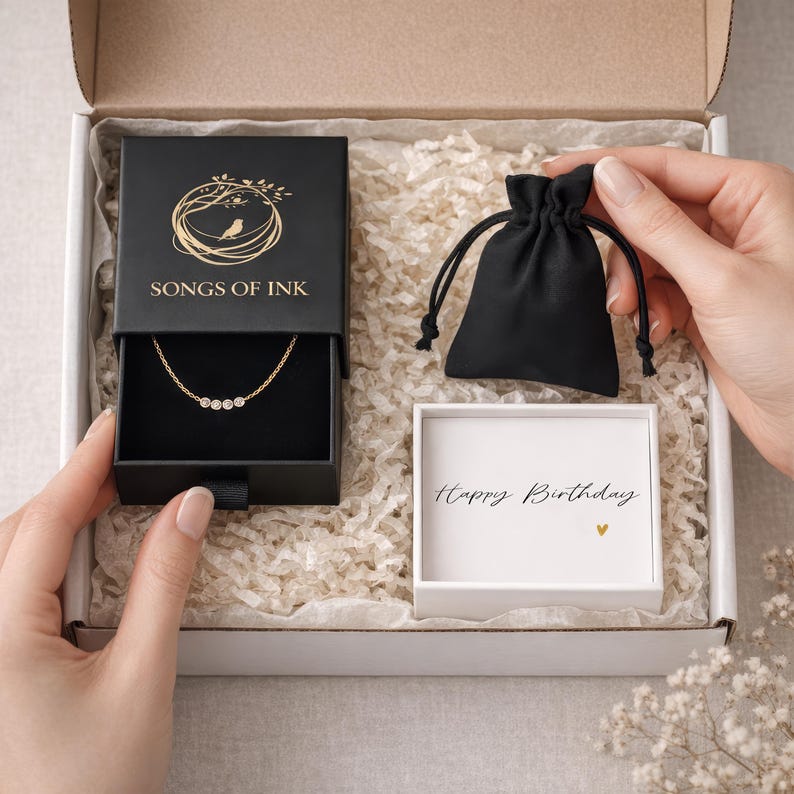 Puede incluir: Una caja de regalo que contiene un collar de oro con tres piedras transparentes, una caja de joyer&iacute;a negra con el texto "SONGS OF INK", una peque&ntilde;a bolsa negra con cord&oacute;n y una caja blanca con "Happy Birthday" escrito. La caja est&aacute; llena de material de embalaje blanco.