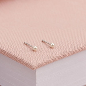 May include: A pair of small stud earrings featuring round white pearls set on silver-toned metal posts. The earrings have a simple design with a smooth, spherical pearl attached to a straight metal pin, creating a classic and understated appearance.