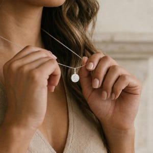 May include: A silver-toned necklace featuring a delicate chain and a small, circular pendant. The pendant has a smooth, reflective surface with a subtle, engraved design visible on its face. The necklace rests against a neutral-coloured garment.