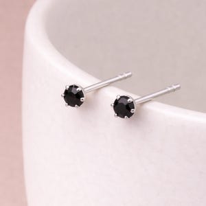 Puede incluir: Un par de pendientes de botón con una pequeña gema negra redonda engastada en metal plateado. Los pendientes se presentan sobre un fondo blanco, destacando su diseño sencillo. Las gemas son el punto focal de la imagen.