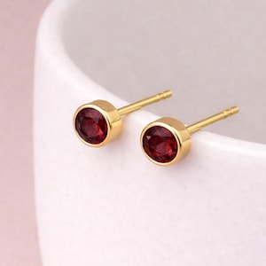 Pode incluir: Um par de brincos de ouro com pedras preciosas redondas de cor vermelha escura. Os brincos têm uma moldura de bisel dourada e um design simples. Os brincos são exibidos em uma superfície branca.