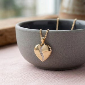 Könnte beinhalten: Ein herzförmiger Medaillon-Anhänger aus Gold, der an einer passenden Goldkette hängt. Das Medaillon hat eine glatte, polierte Goldoberfläche mit einem geschwungenen, vertikalen Kanal, der mit kleinen, klaren, funkelnden Steinen besetzt ist.