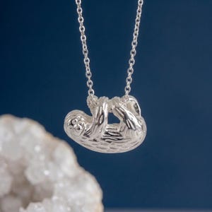 Könnte beinhalten: Ein silberfarbener Anhänger in Form eines Faultiers, das an einem Ast hängt, befestigt an einer zarten silberfarbenen Kette. Die Faultierfigur weist eine texturierte Oberfläche an Körper und Gliedmaßen auf, wobei die Kette an den Vorderpfoten befestigt ist.