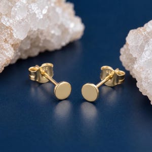以下が含まれることがあります： フラットな円形ディスクデザインのゴールドトーンのスタッドピアス一対。各ピアスには細いポストとバタフライ型のキャッチが付いており、キャッチにはスターリングシルバーを示す925の刻印がはっきりと確認できます。