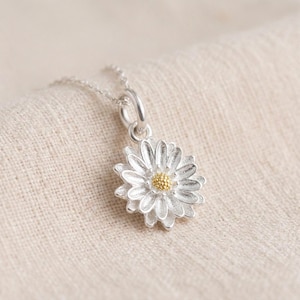 May include: A silver pendant necklace featuring a flower charm with textured petals and a small, round, gold-colored center. The pendant hangs from a delicate silver chain, showcasing a detailed floral design with a metallic finish.