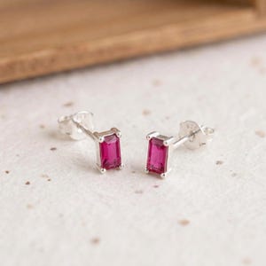 May include: A pair of silver stud earrings featuring rectangular, baguette-cut vibrant pink stones. Each earring is secured with a silver post and a matching butterfly back, showcasing a clean, metallic setting that holds the gemstone firmly.