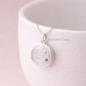 Puede incluir: Un collar con un medall&oacute;n redondo plateado con dise&ntilde;o de estrellas. El medall&oacute;n cuelga de una fina cadena. El texto "Includes Chain" es visible. El medall&oacute;n tiene una superficie texturizada y un cierre.