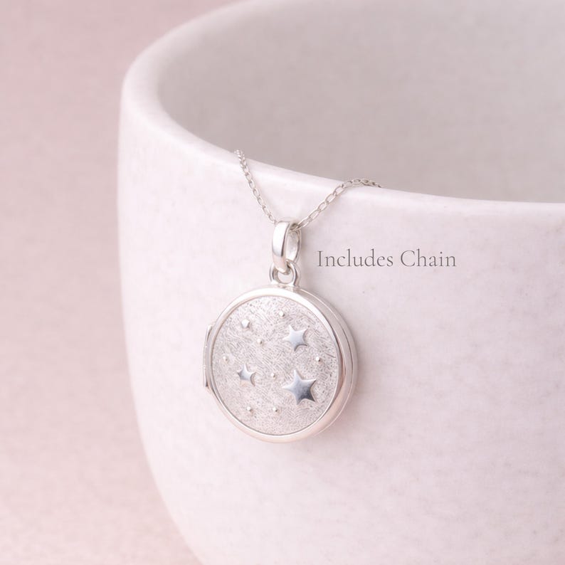 Puede incluir: Un collar con un medall&oacute;n redondo plateado con dise&ntilde;o de estrellas. El medall&oacute;n cuelga de una fina cadena. El texto "Includes Chain" es visible. El medall&oacute;n tiene una superficie texturizada y un cierre.