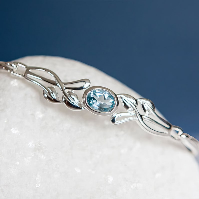 May include: A silver-toned bracelet featuring a central oval-cut light blue gemstone set in a polished metal frame. The band is designed with symmetrical, curved leaf-like metal motifs extending from both sides of the central stone setting.