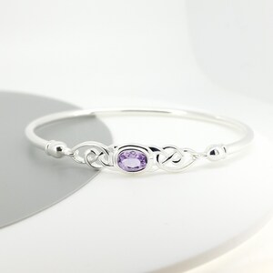 May include: A silver bangle bracelet featuring a central oval-cut purple amethyst gemstone. The metal band displays intricate, interconnected knotwork patterns on both sides of the stone, leading to a smooth, polished silver finish.