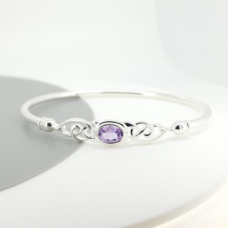 May include: A silver bangle bracelet featuring a central oval-cut purple amethyst gemstone. The metal band displays intricate, interconnected knotwork patterns on both sides of the stone, leading to a smooth, polished silver finish.
