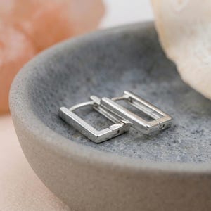 Può includere: Un paio di orecchini a cerchio rettangolari color argento con finitura lucida. Gli orecchini presentano un meccanismo di chiusura a cerniera e un design geometrico pulito. La superficie metallica riflette la luce, mettendo in risalto i bordi.