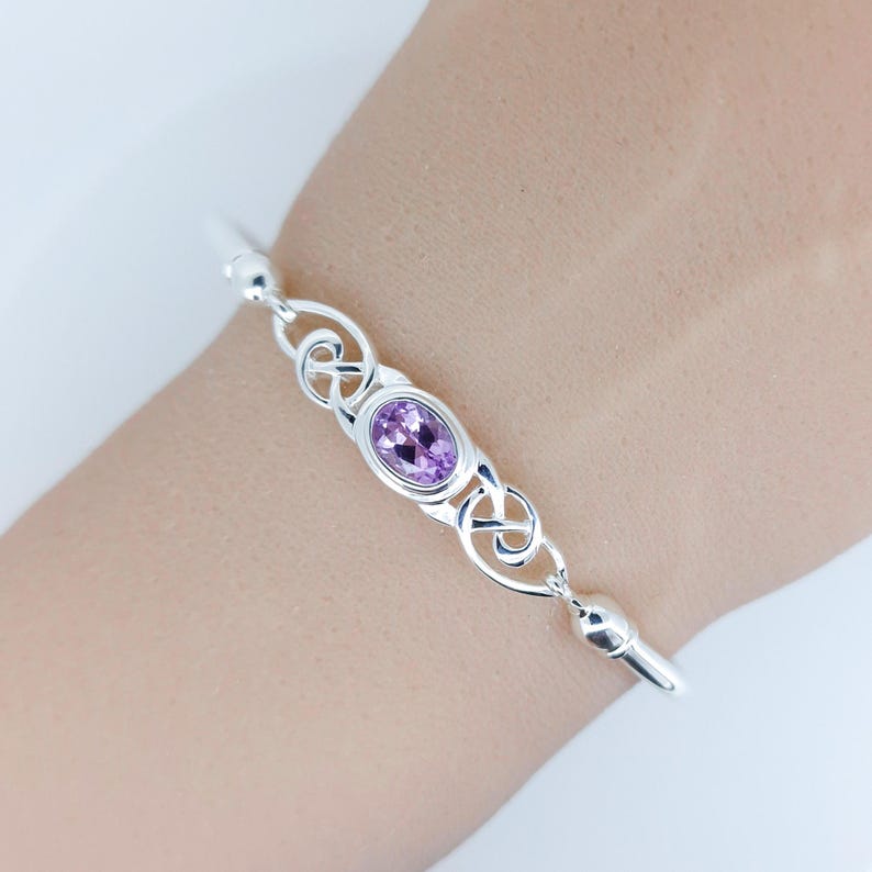 May include: A silver bangle bracelet featuring a central oval-shaped light purple gemstone set in a polished metal frame. The design includes intricate openwork metal loops on both sides of the stone, connecting to a smooth, solid silver band.