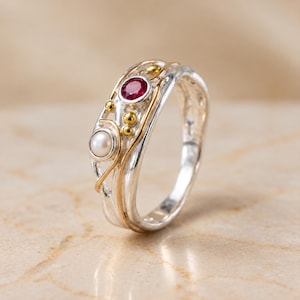 May include: A silver ring featuring a textured, organic band with a thin gold wire accent. The ring is set with one round, deep red ruby and one white pearl, accented by several small, smooth gold spheres embedded along the surface of the band.
