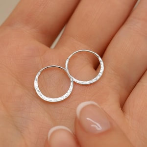 May include: Two small, silver hoop earrings with a hammered texture. The earrings are circular and made of a shiny metal. The earrings are displayed on a light-coloured background.