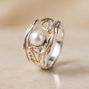 Peut inclure: Une bague large avec une perle blanche centrale sertie dans une monture en argent texturé. Le design comprend de fins fils dorés incurvés qui s'enroulent autour de la perle et sur la surface argentée, créant un aspect organique et superposé.