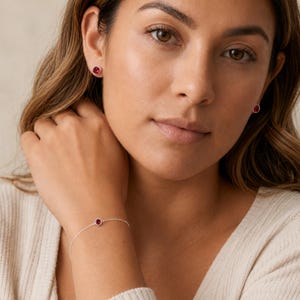 May include: A delicate silver chain bracelet featuring a single round, bezel-set red gemstone at the centre, accompanied by matching round, bezel-set red gemstone stud earrings. The jewellery set displays a consistent, polished metallic finish.