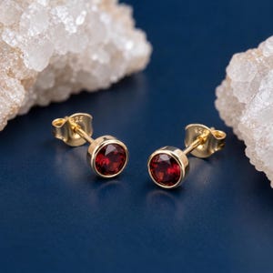 May include: A pair of gold-tone stud earrings featuring round, faceted deep red garnet stones set in a bezel setting. The earring backs are clearly stamped with 925, indicating sterling silver construction with a gold-coloured finish.