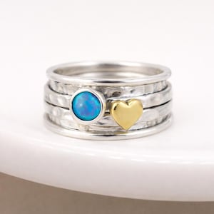Puede incluir: Un anillo de plata con una piedra de ópalo azul y un corazón dorado. El anillo tiene una textura martillada y un diseño apilable. La piedra de ópalo es redonda y está engastada en un bisel de plata. El corazón dorado es liso y pulido.