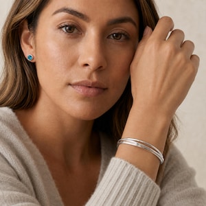 Op de afbeelding: Een set van drie dunne, gepolijste zilverkleurige armbanden om de pols, gecombineerd met een enkele oorsteker met een ronde, felblauwe edelsteen in een zilverkleurige metalen zetting voor een gecoördineerde set.