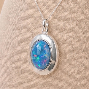Puede incluir: Un collar con colgante de plata que presenta un cabujón de ópalo azul de forma ovalada con motas iridiscentes verdes y rosas. La piedra está engastada en un marco de plata lisa y pulida con una anilla sencilla, suspendido de una cadena fina.