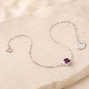 May include: A silver chain bracelet featuring a single round purple gemstone in a bezel setting. The bracelet includes a circular charm engraved with the name abby and a small heart symbol, finished with a spring ring clasp closure.