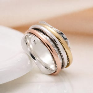 Puede incluir: Un anillo de plata con tres bandas en oro rosa, oro y plata. El anillo tiene un diseño texturizado en la banda dorada y un acabado liso en las otras bandas. El anillo es de talla 19. El anillo está sobre una superficie blanca.