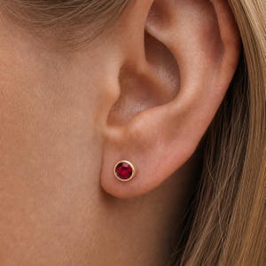 Puede incluir: Primer plano de un pendiente de oro con una gema redonda de color rojo intenso. El pendiente está engastado en un engaste sencillo. El pendiente se muestra en la oreja de una persona.