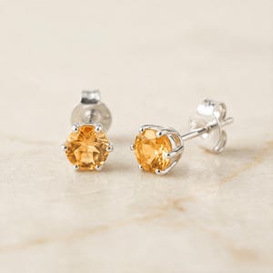 May include: A pair of stud earrings featuring round, faceted, golden-yellow citrine gemstones set in silver-toned metal prongs. Each earring includes a post back mechanism, showcasing the clear, warm-toned stones in a simple, classic design.
