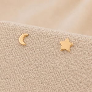 Puede incluir: Un par de pendientes de botón en tono dorado que presentan una forma de media luna y una forma de estrella de cinco puntas. Los pendientes tienen un acabado liso y pulido, diseñados como piezas individuales, incluyendo una luna y una estrella.
