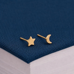 May include: A pair of gold-toned stud earrings featuring a five-pointed star shape and a crescent moon shape. Each earring has a thin metal post extending from the back, designed for pierced ears, with a smooth, polished metallic finish throughout.