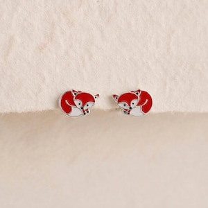 May include: A pair of stud earrings shaped like curled foxes. Each earring features red and white enamel detailing with black accents for the eyes and ears, set against a silver-toned metal outline, creating a small, detailed animal design.