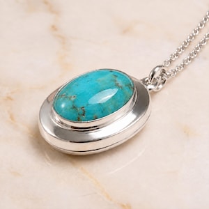 Puede incluir: Un collar con colgante de plata que presenta una piedra preciosa de turquesa lisa y ovalada con vetas naturales de color marrón. La piedra está engastada en un bisel de plata pulida, unido a una delicada cadena de plata con una argolla simple.