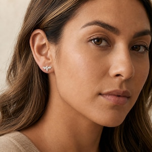 May include: A small, silver-toned stud earring shaped like a bee. The earring features detailed wings and a segmented body, with a polished metallic finish. It is worn on the earlobe, showcasing a compact design that sits flush against the skin.