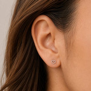 Pode incluir: Um pequeno brinco de pino na cor prateada com o formato de um contorno de coração aberto. O brinco está posicionado na parte inferior do lóbulo da orelha, apresentando uma construção simples de fio metálico fino que forma uma silhueta.