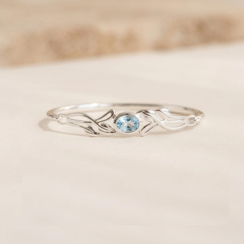 May include: A silver ring featuring a central oval-cut light blue gemstone. The band is designed with delicate, symmetrical metalwork that resembles stylized leaves or vines extending from the center setting on both sides.