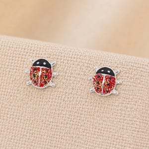 Könnte beinhalten: Ein Paar Ohrstecker in Form von Marienkäfern. Jeder Ohrstecker hat eine silberfarbene Metallumrandung, einen schwarzen Kopf mit zwei weißen Punkten und einen roten Flügelbereich mit goldenen und roten Glitzerakzenten sowie kleinen schwarzen Punkten.