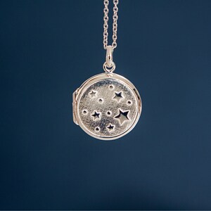 Puede incluir: Collar con colgante de medall&oacute;n plateado. El medall&oacute;n redondo presenta un dise&ntilde;o de estrellas y puntos. La joya se muestra sobre un fondo azul oscuro.