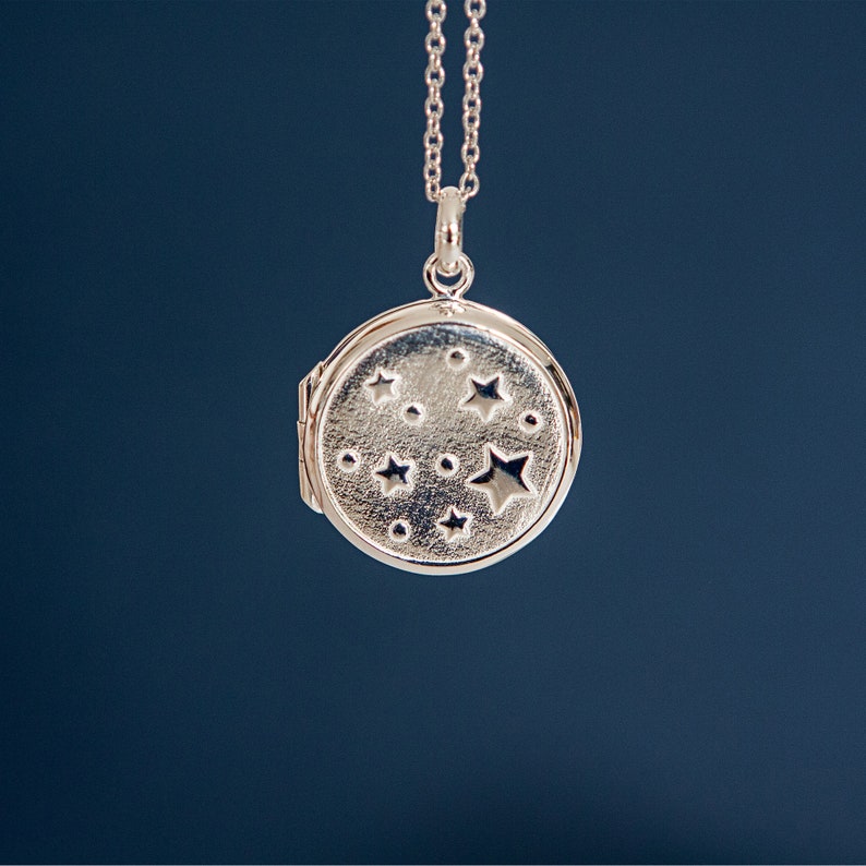 Puede incluir: Collar con colgante de medall&oacute;n plateado. El medall&oacute;n redondo presenta un dise&ntilde;o de estrellas y puntos. La joya se muestra sobre un fondo azul oscuro.