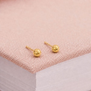 May include: A pair of gold stud earrings featuring small, spherical beads with a textured, faceted surface. Each earring has a thin, straight post extending from the back of the round bead, designed for secure placement in a pierced earlobe.