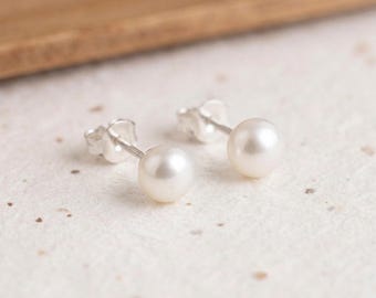 Peut inclure: Une paire de boucles d'oreilles à clous avec des perles blanches rondes montées sur des tiges en argent. Les boucles d'oreilles sont présentées avec la perle vers le haut, révélant la surface lisse et lustrée ainsi que le support en argent.