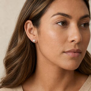 May include: A small, round, silver-coloured stud earring with a textured, dimpled surface. The earring is worn in the earlobe, featuring a metallic finish that catches the light, creating a subtle, reflective appearance against the skin.