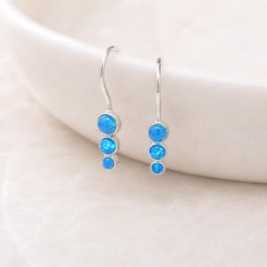 Puede incluir: Un par de pendientes colgantes de plata, cada uno con tres piedras redondas de ópalo azul vibrante. Los pendientes cuelgan de ganchos curvos, mostrando el metal pulido y el color luminoso de los ópalos. Los pendientes se muestran sobre un fondo blanco.