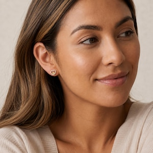 May include: A small stud earring featuring a silver-toned heart outline surrounding a smaller, solid gold-toned heart centre. The earring is worn on the earlobe, showcasing a delicate two-tone metal design with a polished finish and a secure post back.
