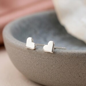 May include: A pair of small, polished silver heart-shaped stud earrings. Each earring features a smooth, reflective surface in the shape of a classic heart, complete with a thin metal post extending from the back for secure attachment to the ear.